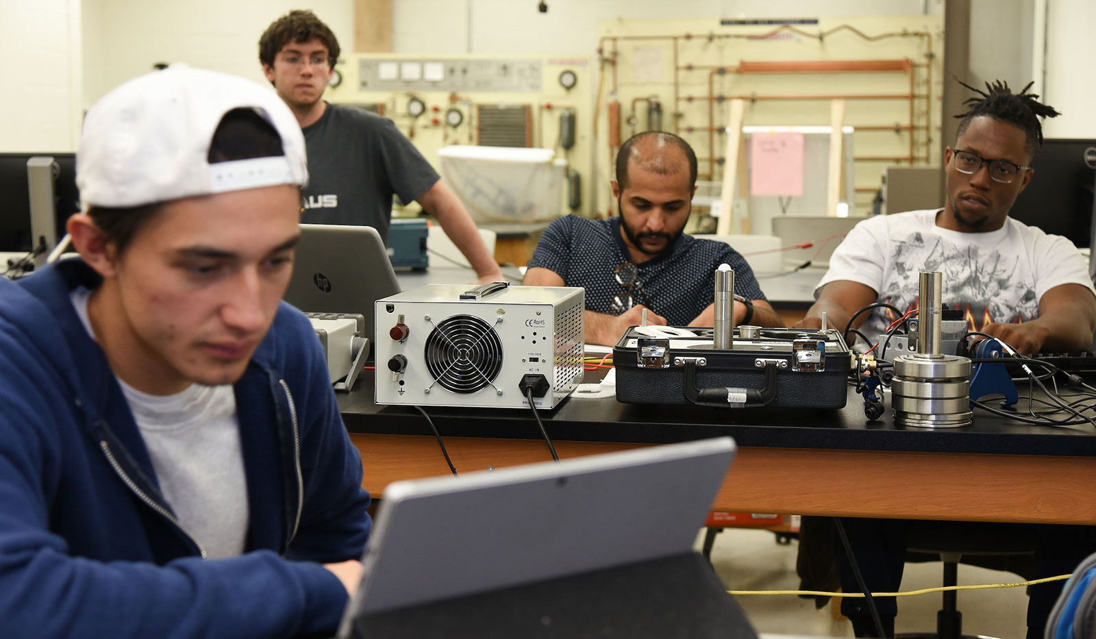 Four male students in engineering class with various mechanical equipment on the table in front of them.